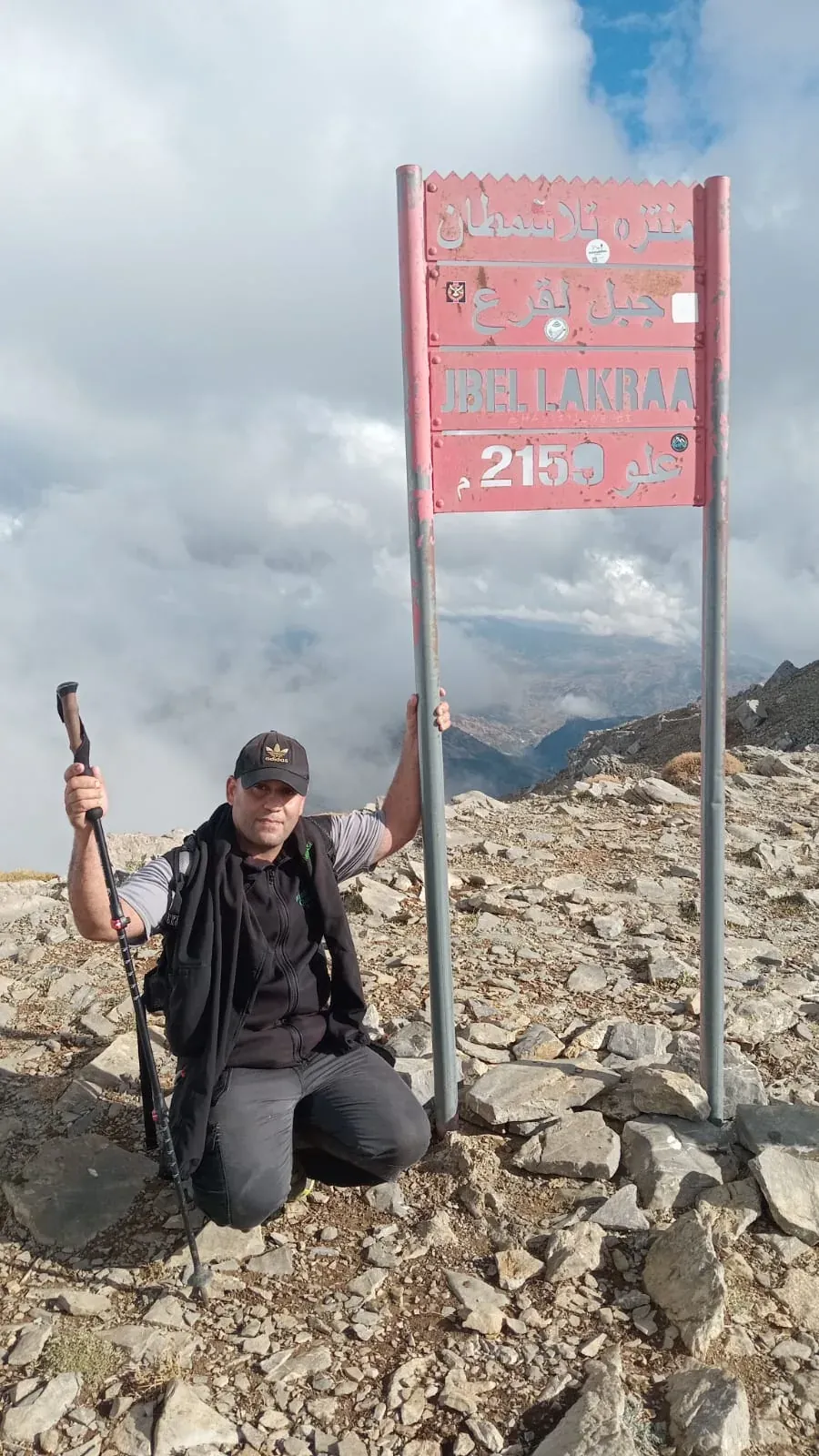 Mountain guide Imad Ben Yahya by the summit sign of Jbel Lakraa, 2159 m, with clouds around the mountains